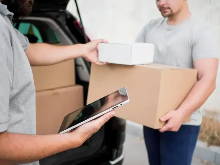 A imagem mostra um homem entregando caixas em um carro, enquanto outra pessoa filma o processo com um smartphone. O cenário sugere uma operação de entrega de produtos, possivelmente para um comércio eletrônico ou um serviço de drive-to-store. No contexto do artigo "Drive-to-store: por que este conceito merece atenção?", a imagem pode ser usada para ilustrar o conceito de drive-to-store, que envolve a entrega de produtos diretamente no veículo do cliente ou em um local de sua escolha, como um estacionamento ou um drive-through.