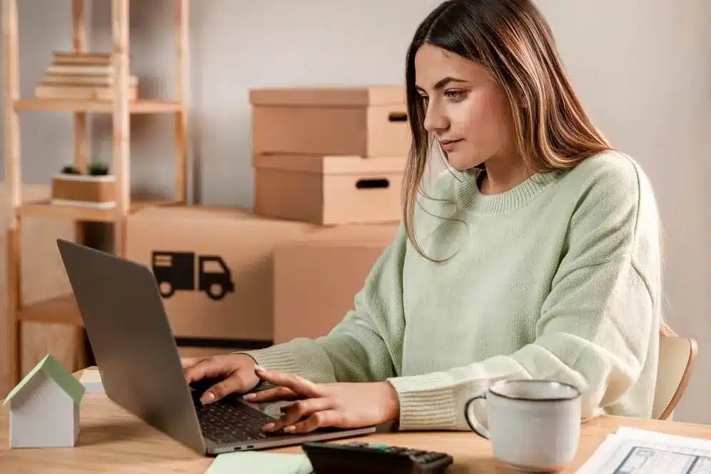 A imagem mostra uma mulher trabalhando em um laptop em um ambiente de home office, com caixas de entrega ao seu redor, representando o crescimento do comércio online. No contexto do artigo, isso simboliza o novo normal dos negócios on-line, onde a adaptação a modelos de trabalho remoto, a importância da logística e a personalização da experiência do cliente estão se tornando padrões para o sucesso no varejo digital.