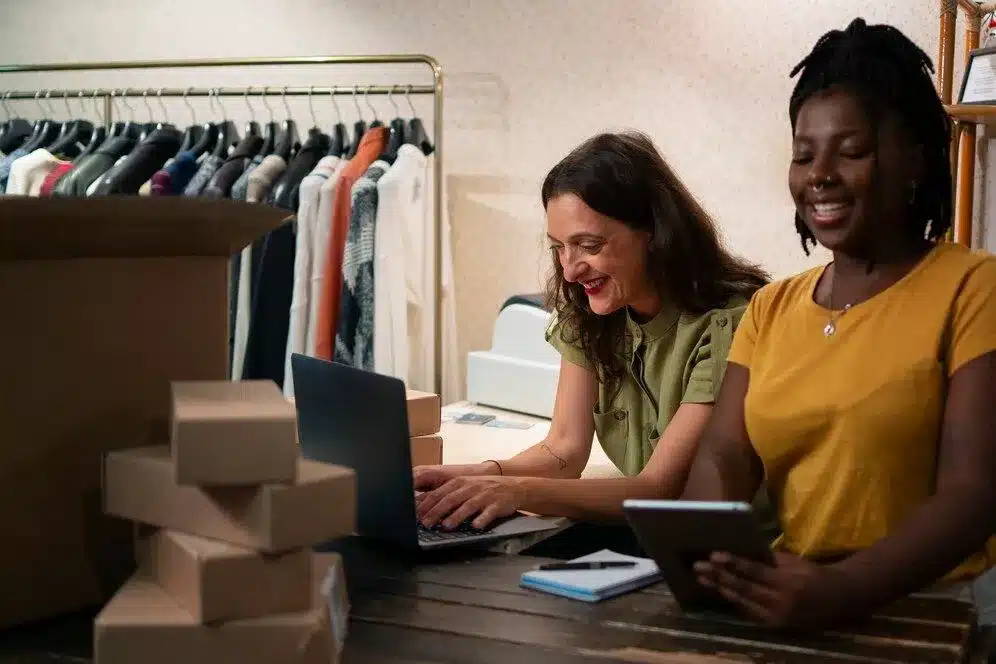A imagem mostra duas mulheres, uma caucasiana e outra afro-americana, trabalhando juntas em um ambiente de loja de varejo. Elas estão usando computadores e analisando informações, provavelmente relacionadas a vendas e gestão de estoque. Essa imagem é relevante para o artigo "O que você precisa saber sobre arquitetura omnichannel para e-commerce" porque ela ilustra a integração de diferentes canais e a colaboração entre equipes em uma estratégia omnichannel.