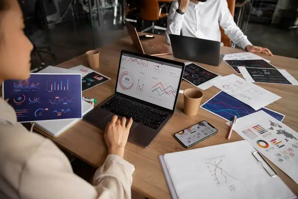 A imagem mostra uma reunião de negócios com colaboradores analisando gráficos e relatórios, simbolizando a gestão de informações e processos. No contexto do artigo sobre RPA (Automação de Robótica Processual), isso reflete como a implementação da RPA pode aumentar a produtividade, ao automatizar tarefas repetitivas e permitir que os funcionários se concentrem em atividades mais estratégicas e criativas, resultando em uma gestão mais eficiente das operações da empresa.