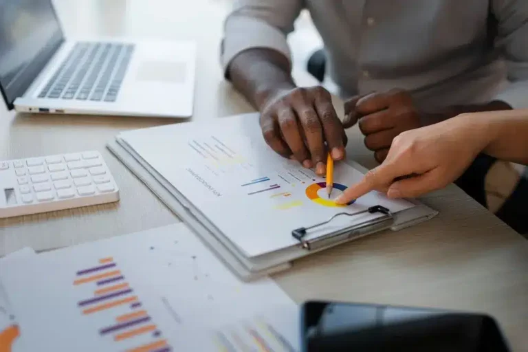 A imagem mostra uma pessoa analisando relatórios financeiros ou de negócios em uma folha de papel, enquanto outro indivíduo aponta para a folha. O ambiente é um escritório com um laptop e um teclado, o que indica um ambiente de trabalho corporativo. A pessoa que está analisando os relatórios está usando uma caneta para apontar detalhes nos gráficos. No contexto do artigo "Como os relatórios inteligentes facilitam a gestão do seu negócio", a imagem pode ser usada para ilustrar como a tecnologia e os relatórios inteligentes podem simplificar e melhorar a análise de dados para tomada de decisão.