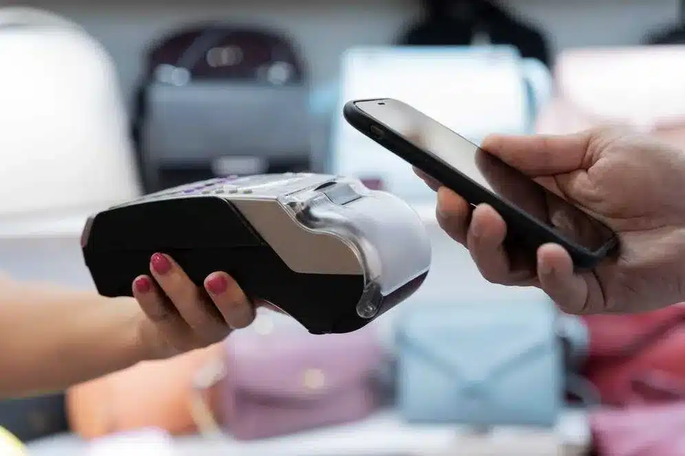 A imagem mostra uma pessoa realizando um pagamento com um smartphone, utilizando um leitor de cartão, representando a modernização e a simplicidade do processo de pagamento. No contexto do artigo sobre o PIX, isso ilustra como essa nova forma de pagamento digital, oferecida pelo Banco Central do Brasil, está facilitando transações rápidas e seguras no dia a dia dos usuários, permitindo operações financeiras instantâneas entre diferentes instituições financeiras.