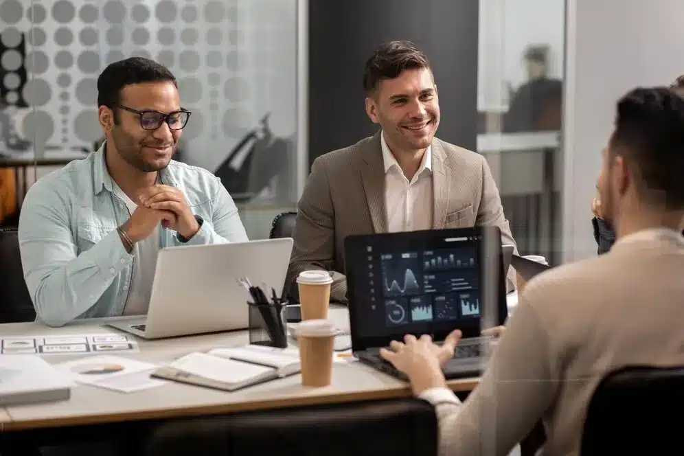 A imagem mostra um grupo de profissionais reunidos em uma reunião de trabalho, discutindo e analisando dados apresentados em um tablet. Ilustra a importância de antecipar as tendências em gestão, pois a equipe parece estar envolvida em um processo de análise e discussão de informações que podem subsidiar decisões estratégicas e melhorar a eficácia da gestão.