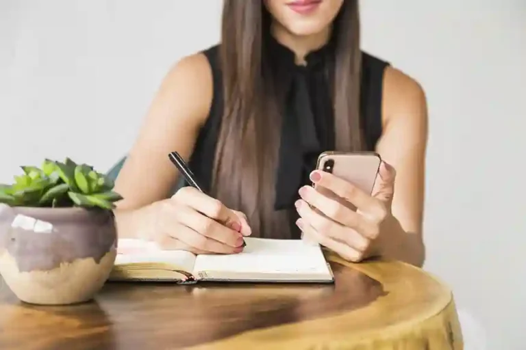A imagem mostra uma mulher escrevendo em um caderno enquanto segura um smartphone. Ela está sentada em uma mesa de madeira com uma planta em vaso ao lado. A imagem sugere que o artigo pode abordar o tema de reserva de emergência para empresas, orientando sobre como as empresas podem se preparar e manter uma reserva financeira para situações inesperadas.
