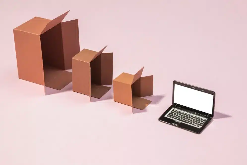 A imagem apresenta um notebook com tela em branco e três caixas de papelão de diferentes tamanhos, dispostas de forma crescente, do menor para o maior, ao lado do notebook. As caixas estão abertas e vazias, e o notebook está ligado, mas sem exibir nenhum conteúdo específico. No contexto do artigo "Módulos ERP: o que são e para que servem?", a imagem pode ser usada para ilustrar a ideia de que os módulos ERP são componentes distintos e escaláveis de um sistema de planejamento empresarial, similarmente às caixas de diferentes tamanhos.