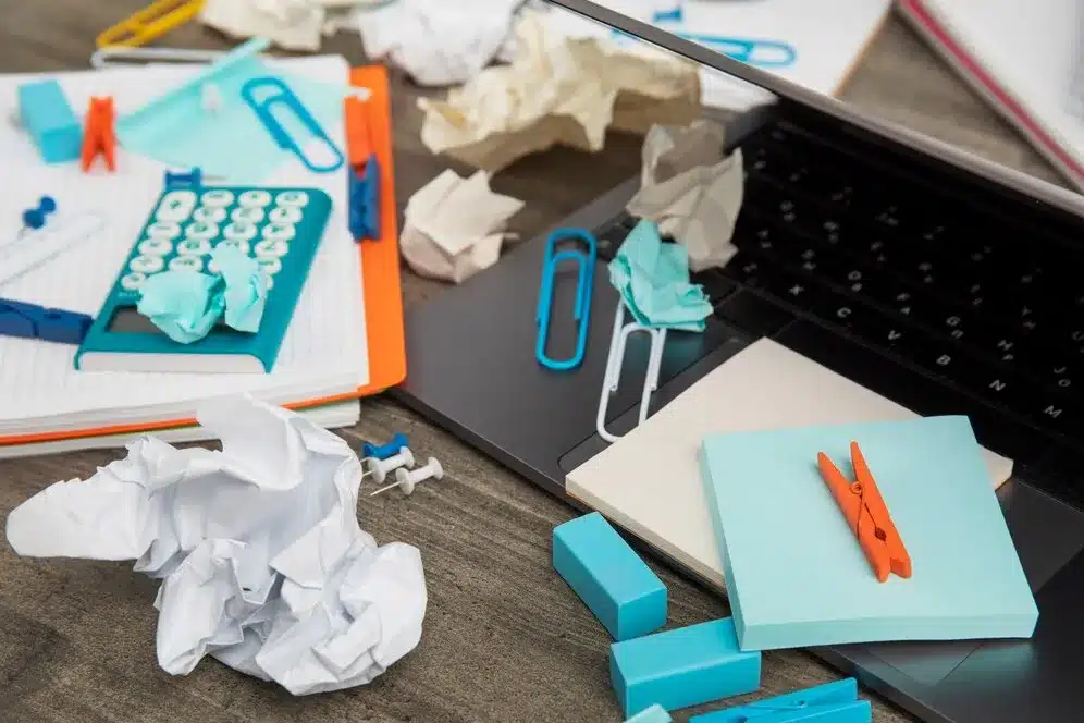 A imagem mostra uma mesa de trabalho desorganizada com um laptop, papel crumpled (papel amassado), blocos de notas, clipes de papel, uma calculadora e outros itens espalhados de forma caótica. Essa cena visualiza um ambiente de trabalho sem ordem, o que pode ser um reflexo de um processo de trabalho ou gestão desordenada.