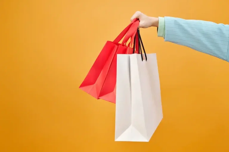 Mão segurando duas sacolas de compras, uma vermelha e uma branca, em um fundo amarelo. Representa a experiência do cliente e a importância de adicionar valor ao seu negócio através de experiências de compra satisfatórias e personalizadas.