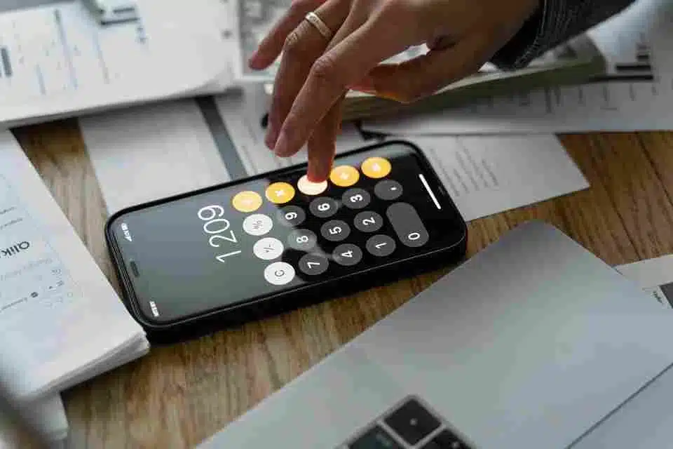 Mão usando um calculadora digital em um smartphone, com documentos de contabilidade ao seu redor. Representa a importância do DANFE (Documento Auxiliar de Notas e de Feituras Eletrônicas) para os negócios, refletindo a necessidade de gerenciamento financeiro e contabilidade digital.