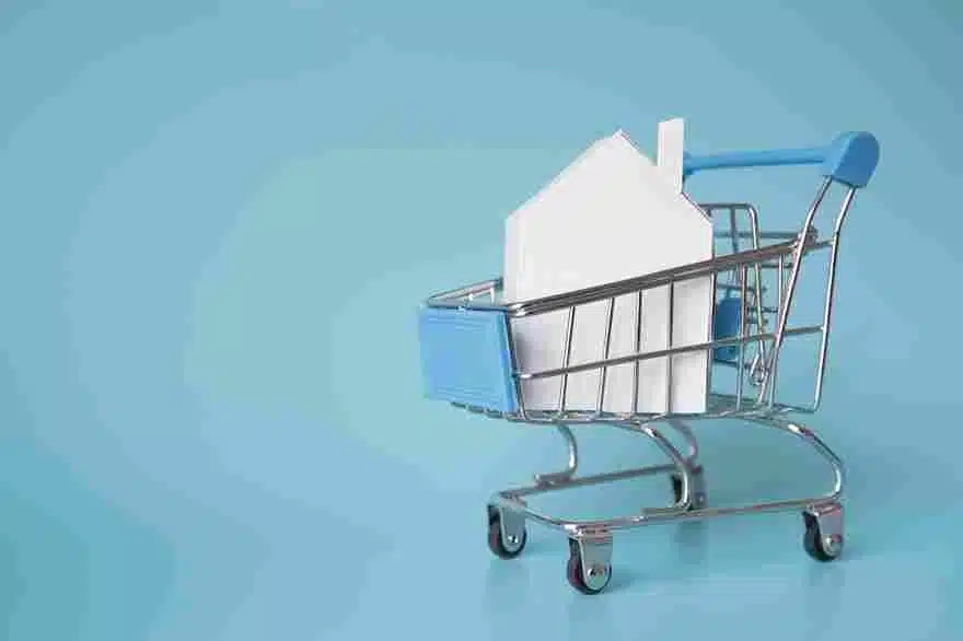 Carrinho de compras com uma casa de papel, simbolizando a compra de imóveis online ou a ideia de "comprar" um lar. Representa a tendência de compras digitais e a integração do comércio imobiliário ao mundo digital.