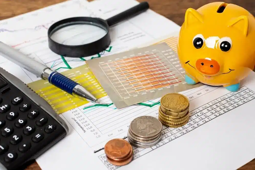 Imagem exibe um ambiente de trabalho financeiro, com um cofre de porco, moedas, uma lupa, um pen, um calculator e papéis com gráficos e tabelas, representando a gestão de finanças e contabilidade. No contexto do artigo sobre mudanças na EFD-Reinf (Declaração de Informações de Contribuintes com Regime de Tributação Substitutiva), isso reflete a importância de estar atualizado com as novas regras e requisitos fiscais, para que os negócios possam cumprir adequadamente suas obrigações e aproveitar ao máximo as facilidades oferidas por essa modalidade tributária.