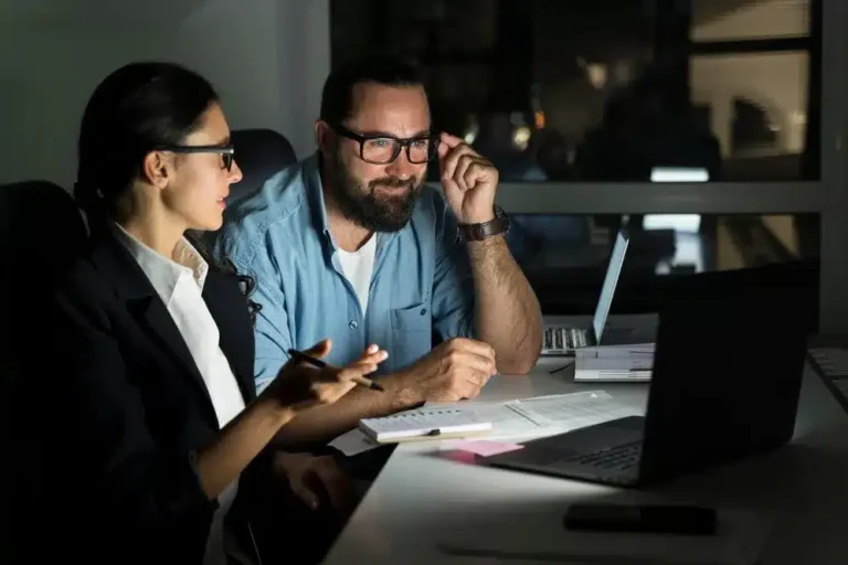 A imagem mostra um homem e uma mulher em um ambiente de trabalho, provavelmente em uma reunião ou discussão sobre negócios. O homem, que parece estar na posição de liderança ou consultor, está apontando para um documento e discutindo com a mulher, que está tomando notas. Ambos estão olhando para um laptop que está em frente a eles, o que indica que eles podem estar analisando dados ou informações relevantes ao ERP (Sistema de Planejamento Empresarial).