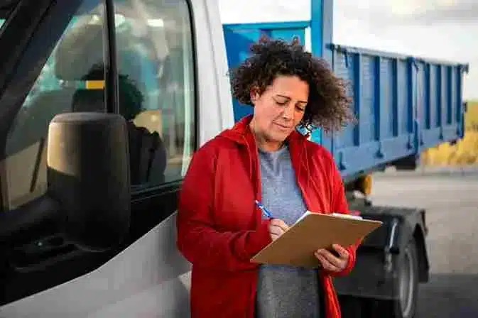 A imagem mostra uma mulher anotando informações ao lado de um caminhão, simbolizando a logística de transporte no contexto do comércio eletrônico. No artigo, isso reflete os principais desafios do transporte logístico, como a necessidade de gerenciar a cadeia de suprimentos eficientemente, garantir a entrega rápida e segura dos produtos, e lidar com questões de custo e regulamentação, para garantir a satisfação dos clientes e a rentabilidade do negócio.