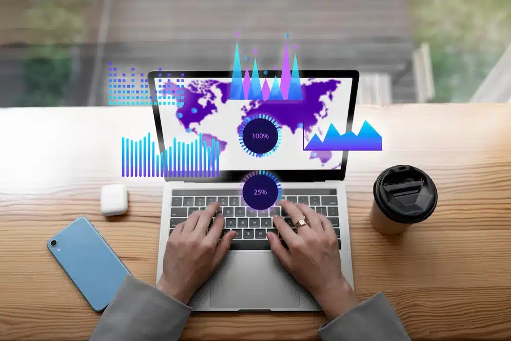 A imagem mostra uma pessoa sentada em uma mesa, usando um laptop que exibe gráficos e mapas interativos, o que sugere análise de dados e monitoramento em tempo real. Ao lado do laptop, há um smartphone e uma taza de café, indicando um ambiente de trabalho moderno e conectado.