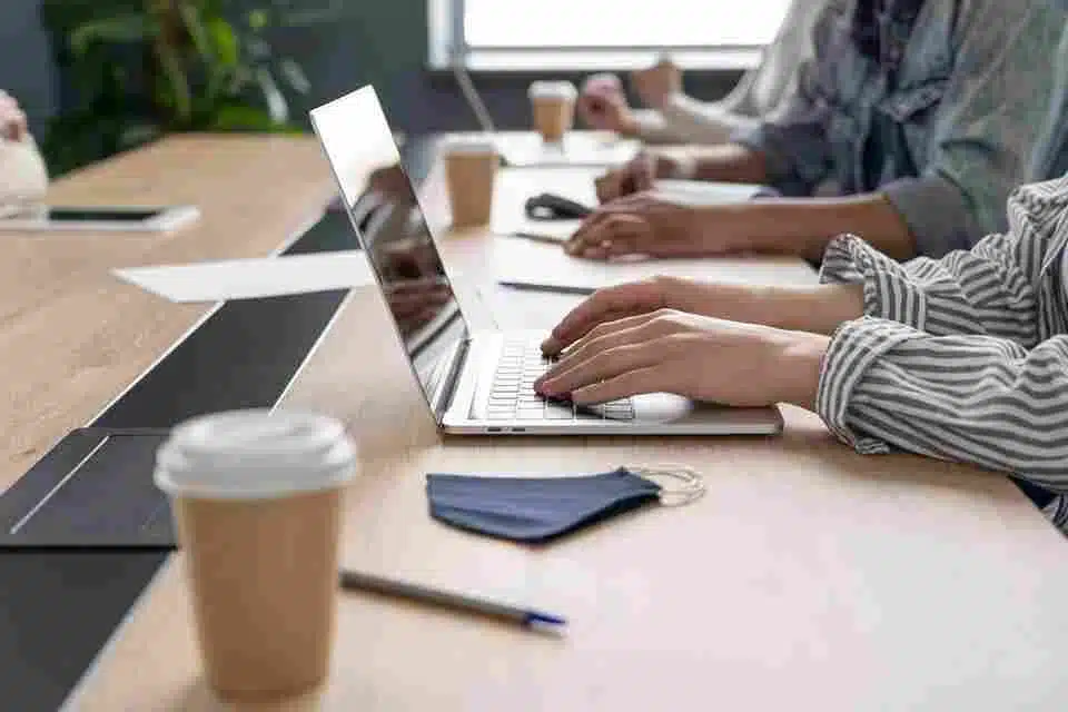 A imagem mostra um ambiente de trabalho com várias pessoas usando computadores, representando um cenário típico de backoffice de vendas no varejo. Os participantes estão focados em suas tarefas, com lembretes e café na mesa, sugerindo um ambiente de colaboração e gerenciamento de operações diárias, que é crucial para o backoffice de vendas em lojas de varejo.