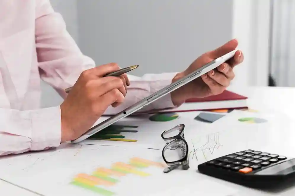 A imagem mostra uma pessoa em um ambiente de trabalho, usando uma caneta para anotar algo em uma folha de papel, enquanto a outra mão segura um smartphone. Em sua frente, há um calculadora, um par de óculos, e uma folha com gráficos coloridos, sugerindo que ela está envolvida em atividades de análise de dados ou gestão financeira.
