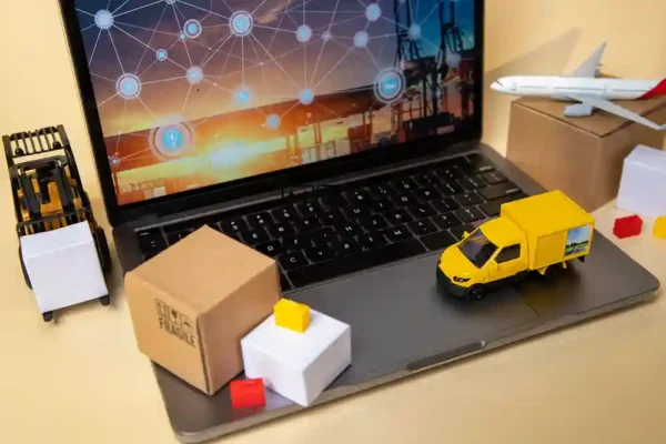 A imagem mostra a representação de uma cadeia de suprimentos digital e logística, com foco em tecnologia. Um laptop, com uma tela exibindo uma rede de conexões e ícones de transporte, serve como base. Sobre o teclado e ao lado do laptop, há miniaturas que simbolizam o fluxo de mercadorias: um caminhão de entrega amarelo, uma empilhadeira (forklift), e um avião de carga posicionado sobre uma caixa de papelão. As caixas de transporte (uma marrom com símbolos de "frágil" e uma branca) completam a cena. O conjunto ilustra a integração de logística, e-commerce, e transporte impulsionada pela tecnologia e pela gestão digital.