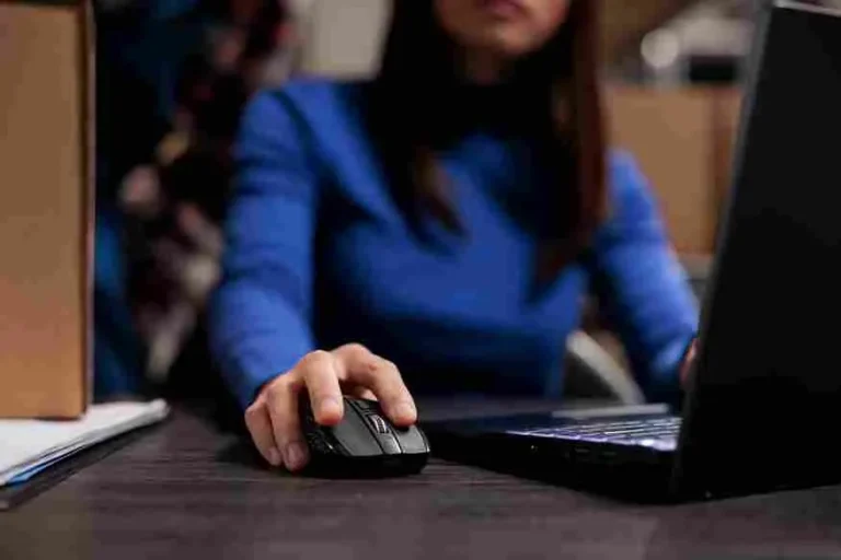 Close-up das mãos de uma mulher utilizando um mouse de computador em uma mesa de escritório escura. Ela veste uma blusa azul vibrante. Ao fundo, caixas de papelão sugerem um ambiente de expedição ou estoque. A imagem ilustra o trabalho técnico de configuração tributária e análise de dados para o cálculo do IPI na base de cálculo do ICMS, focando em precisão contábil e gestão de e-commerce B2B.