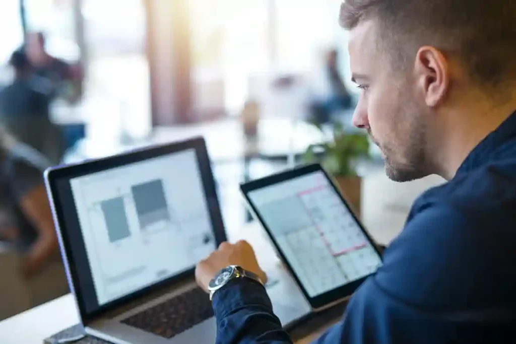Homem jovem em ambiente de escritório moderno utiliza um notebook e um tablet simultaneamente para monitorar dashboards de um sistema de gestão empresarial (ERP). A imagem destaca a mobilidade e a integração de dados em tempo real, mostrando gráficos e indicadores de desempenho. Ideal para ilustrar tecnologia de gestão, automação de processos e produtividade corporativa.