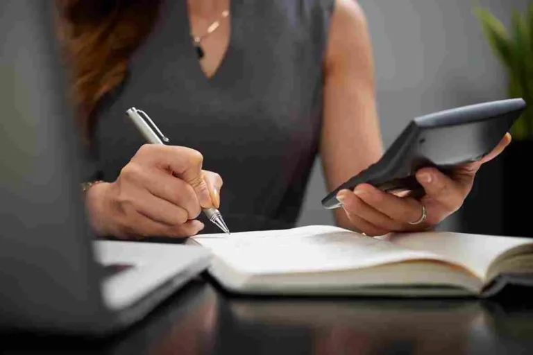 Visão aproximada das mãos de uma mulher em traje executivo cinza, escrevendo em um caderno de anotações enquanto segura uma calculadora financeira. Ao lado, um notebook aberto sugere um ambiente de análise contábil. A imagem ilustra o conceito de Responsabilidade Tributária e a gestão de riscos fiscais em 2026, focando em precisão, conformidade e auditoria financeira empresarial.
