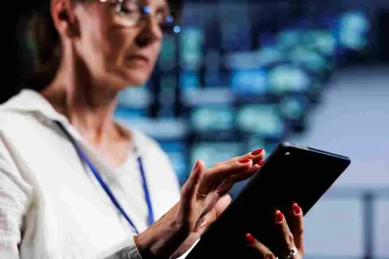 Mulher com óculos e traje profissional opera um tablet em um ambiente de alta tecnologia com múltiplos monitores ao fundo exibindo dashboards de segurança e vigilância. A imagem ilustra o uso de tecnologia antiperdas e inteligência artificial no varejo alimentar para monitoramento de inventário, prevenção de furtos e controle de quebras operacionais em tempo real.
