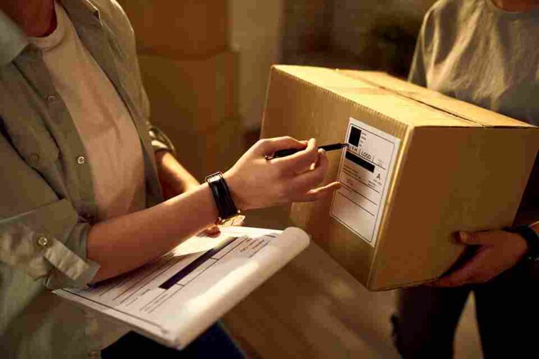Visão aproximada de dois profissionais em um centro de distribuição. Um deles segura uma prancheta com relatórios de conferência enquanto aponta com uma caneta para a etiqueta de uma caixa de papelão segurada pelo outro colega. A imagem ilustra a etapa crítica de expedição, conferência de pedidos e gestão logística de uma distribuidora, destacando a eficiência operacional e o controle de fluxo de saída