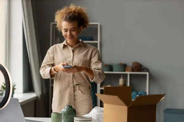 Jovem empreendedora sorridente vestindo camisa bege, operando um smartphone em um estúdio ou escritório aconchegante. À sua frente, uma caixa de papelão aberta e produtos de decoração sugerem o processo de preparação de pedidos. A imagem ilustra o papel de um integrador de marketplace na automação de vendas, permitindo a gestão centralizada de estoque, pedidos e expedição de diversos canais de venda em uma única plataforma, otimizando o fluxo operacional para e-commerces que buscam escala.
