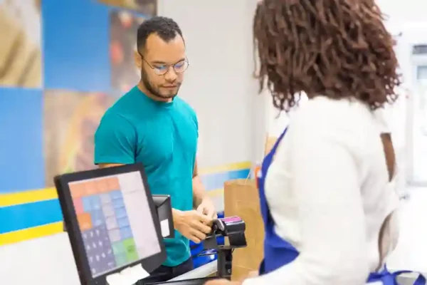 Visão de um cliente homem, vestindo camiseta verde e óculos, realizando o pagamento em um ponto de venda (PDV) de uma loja física. Ele interage com uma funcionária que opera o sistema de caixa, enquanto uma tela sensível ao toque exibe a interface do sistema de vendas. A imagem ilustra o conceito de PDV (Ponto de Venda) em funcionamento, destacando a agilidade no atendimento e a integração necessária com um sistema de gestão para controle de estoque, emissão de nota fiscal e automação comercial.
