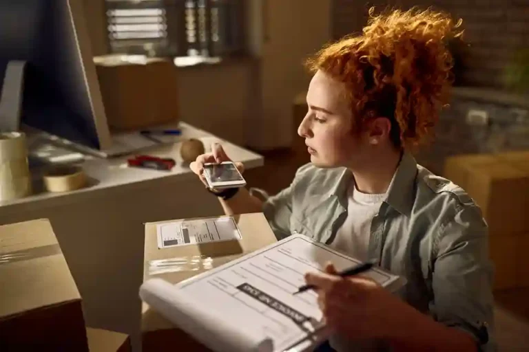 Jovem mulher ruiva com cabelo preso em um coque trabalha no setor de expedição e faturamento de uma empresa à noite. Ela segura uma prancheta com relatórios fiscais enquanto utiliza um smartphone para escanear a etiqueta de identificação ou QR Code de uma caixa de papelão pronta para envio. Ao redor, há caixas embaladas e fita adesiva sobre a bancada. A imagem ilustra o processo de expedição logística e a conferência necessária para a inclusão do frete na base de cálculo do ICMS e do ICMS-ST em 2026.