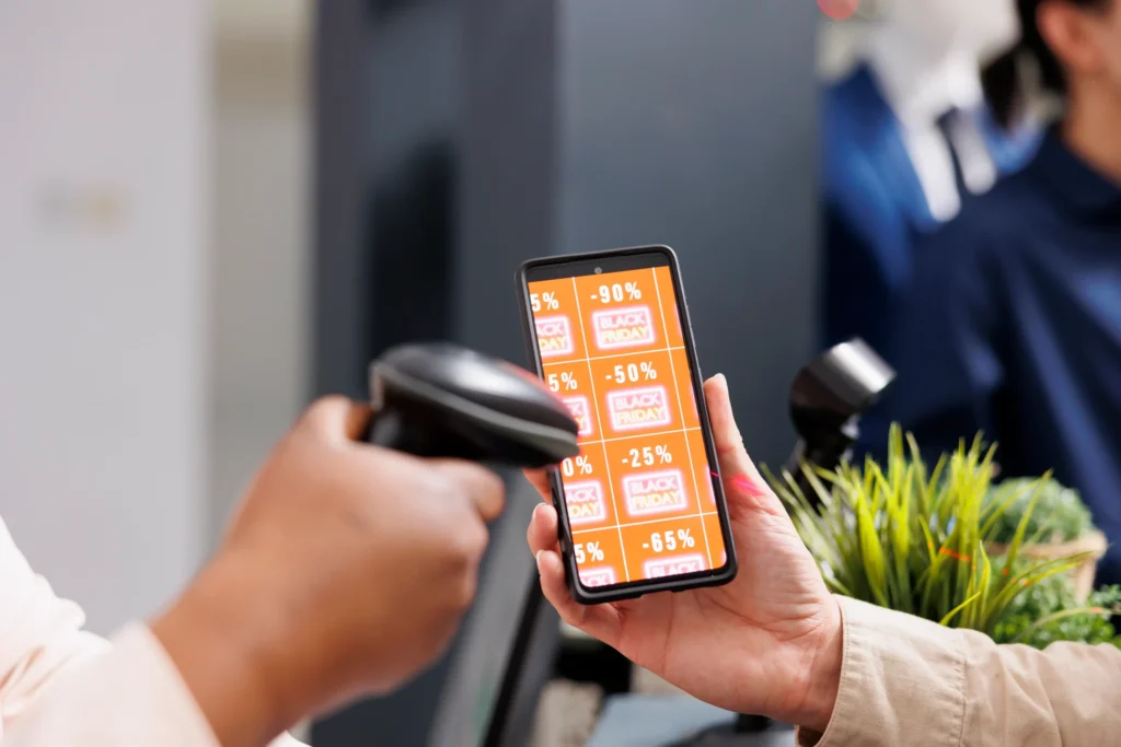 Close-up das mãos de uma pessoa segurando um smartphone que exibe uma grade de cupons de desconto de Black Friday, com ofertas variando de 25% a 90%. Outra pessoa utiliza um leitor de código de barras para digitalizar o dispositivo em um ponto de venda. A imagem ilustra a integração entre e-commerce e loja física, destacando a importância de um sistema de gestão empresarial (ERP) robusto para gerenciar promoções omnichannel, estoque e precificação dinâmica com segurança.