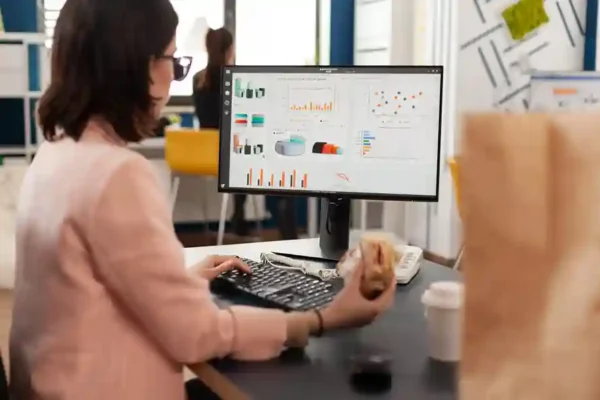 Visão lateral de uma mulher trabalhando sentada em uma mesa de escritório moderna. Ela usa óculos, blazer rosa claro e digita em um teclado preto enquanto analisa gráficos analíticos e relatórios de desempenho em um grande monitor de computador. Na mesa, há um copo de café descartável e uma embalagem de papel pardo desfocada em primeiro plano. A imagem ilustra a centralização de dados, análise de KPIs e o uso de um sistema ERP para e-commerce da ONCLICK para otimizar operações de vendas online.