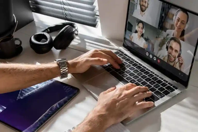 Visão de cima das mãos de um profissional digitando no teclado de um notebook aberto em uma mesa de home office. A tela do computador exibe uma videoconferência ativa com quatro colegas de equipe em chamada de vídeo. Ao lado, há um tablet desligado com reflexos na tela, um headphone preto de ouvido e uma caneca de café perto de uma janela com persianas. A imagem retrata o cenário de um trabalho remoto desorganizado, ilustrando os desafios de comunicação, falta de integração de processos internos e falhas na cultura corporativa digital.