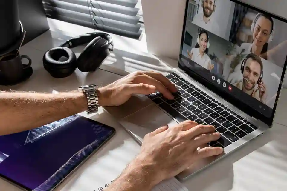 Visão de cima das mãos de um profissional digitando no teclado de um notebook aberto em uma mesa de home office. A tela do computador exibe uma videoconferência ativa com quatro colegas de equipe em chamada de vídeo. Ao lado, há um tablet desligado com reflexos na tela, um headphone preto de ouvido e uma caneca de café perto de uma janela com persianas. A imagem retrata o cenário de um trabalho remoto desorganizado, ilustrando os desafios de comunicação, falta de integração de processos internos e falhas na cultura corporativa digital.